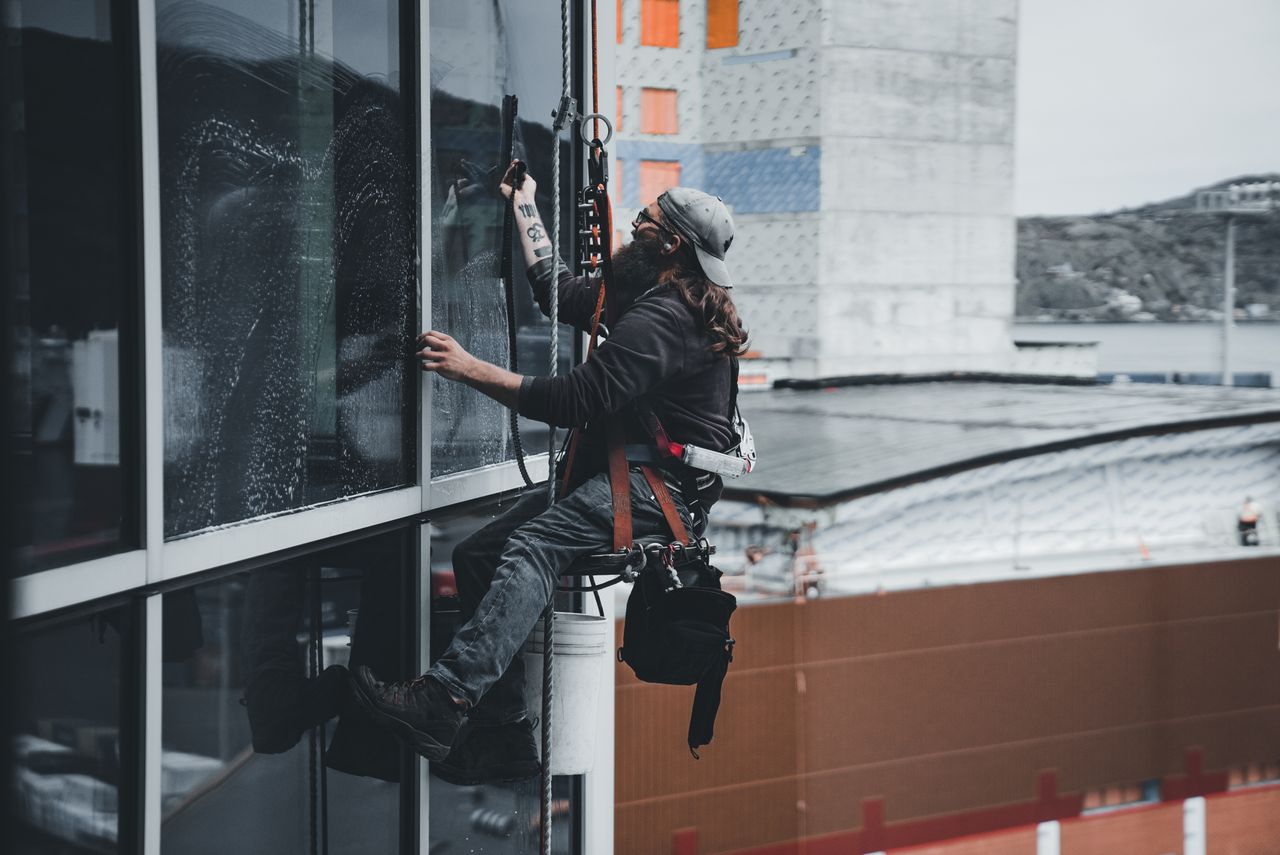 Un homme nettoie les vitres d’un gratte-ciel, harnais de sécurité en place, illustrant un travail en hauteur en milieu urbain.